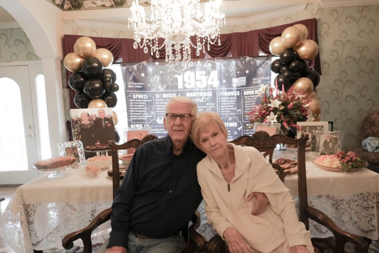 An elderly couple sitting together in a decorated room with a chandelier and party setup.