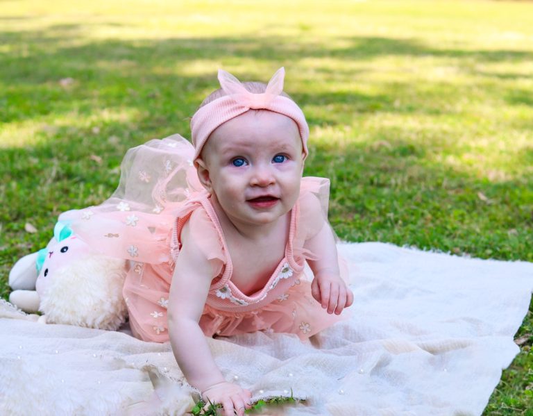 A smiling baby in a pink dress crawls on a blanket in a grassy area.