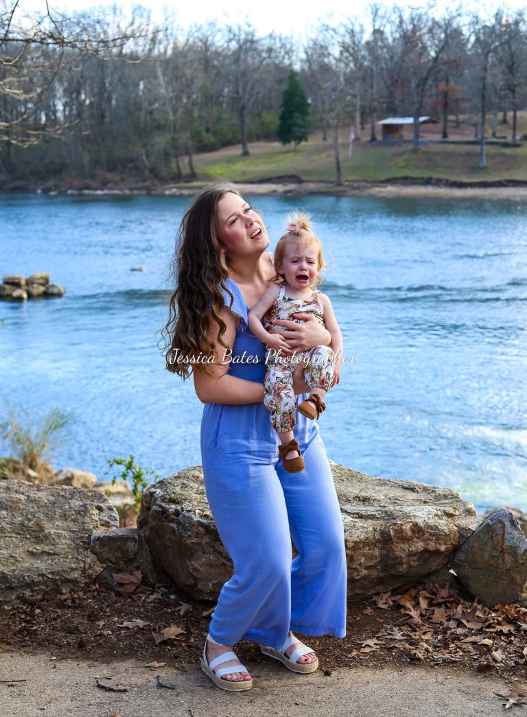 A woman in a blue outfit holds a crying toddler by a river.