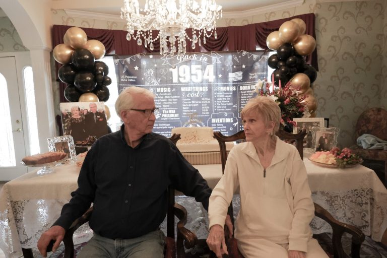 An elderly couple sitting together in a decorated room with balloons and a banner.